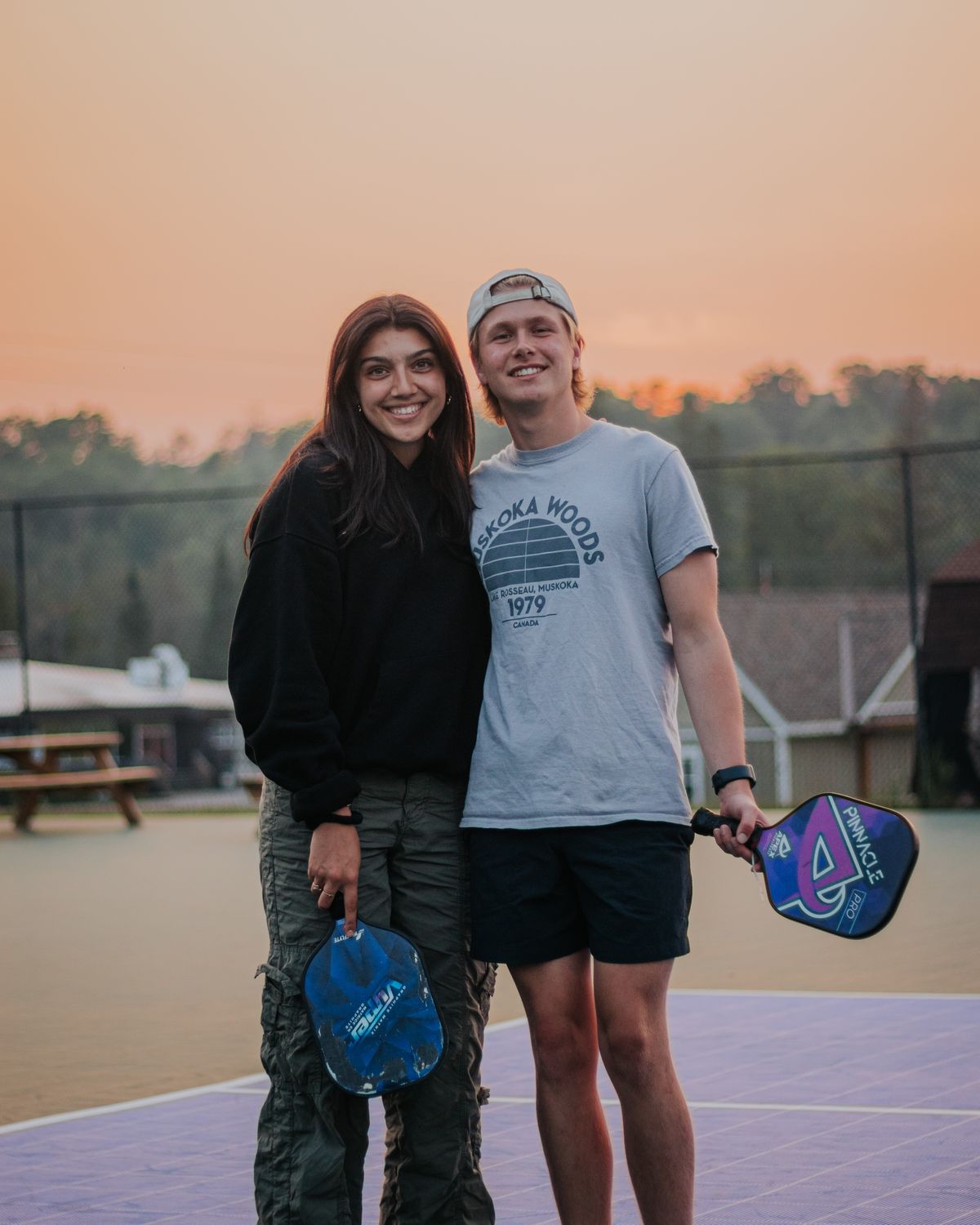 Two friends with pickleball paddles on an outdoor court at sunset.
