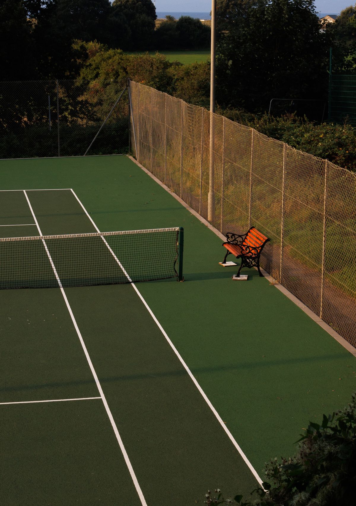 An empty tennis court and bench in warm golden-hour light.