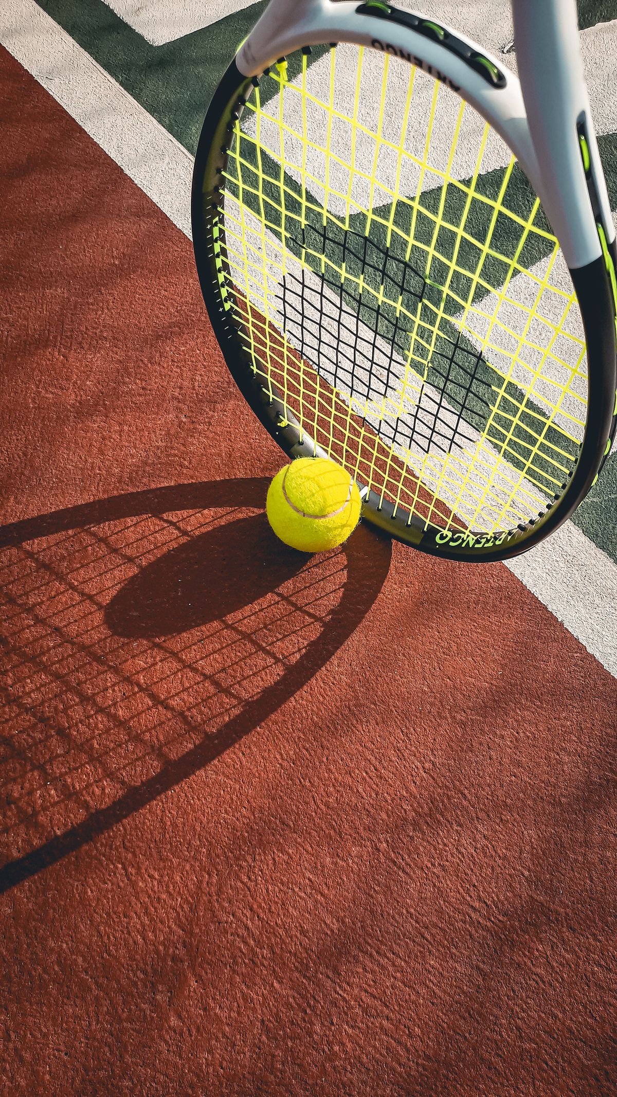 A tennis racket and yellow ball casting long shadows across a sunlit court.